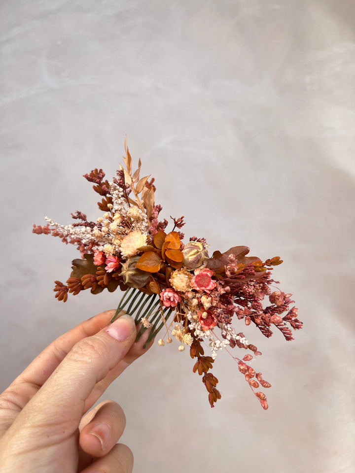 A hand holding a dried floral hair comb featuring autumnal shades of rust, terracotta, peach, and cream against a soft, textured light gray background.