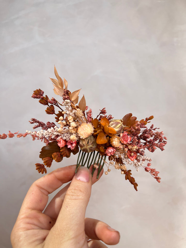 A hand holding a small dried floral hair comb accessory with autumnal shades of brown, terracotta, peach, and cream against a soft light gray background.