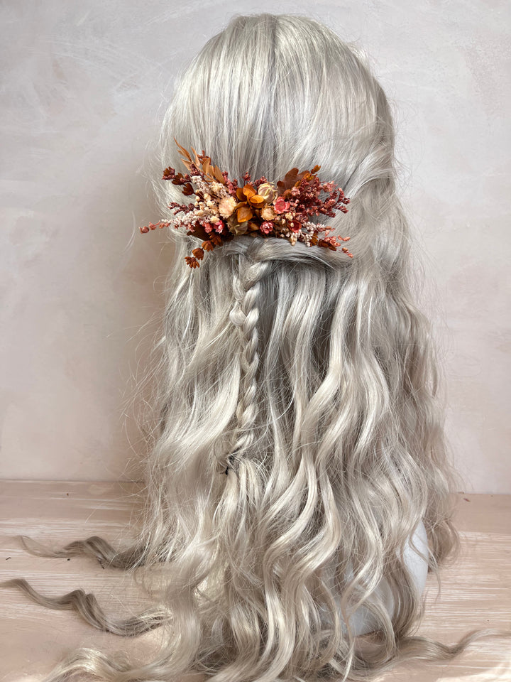 Long wavy silver-gray wig with a half-up style, side braid, and a decorative floral hair accessory in orange, rust, and brown tones, shown against a soft neutral background.