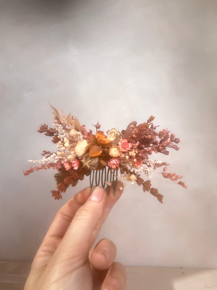 A dried floral hair comb accessory held by a hand, featuring muted autumn tones of terracotta, dusty rose, beige, and brown against a soft gray background.