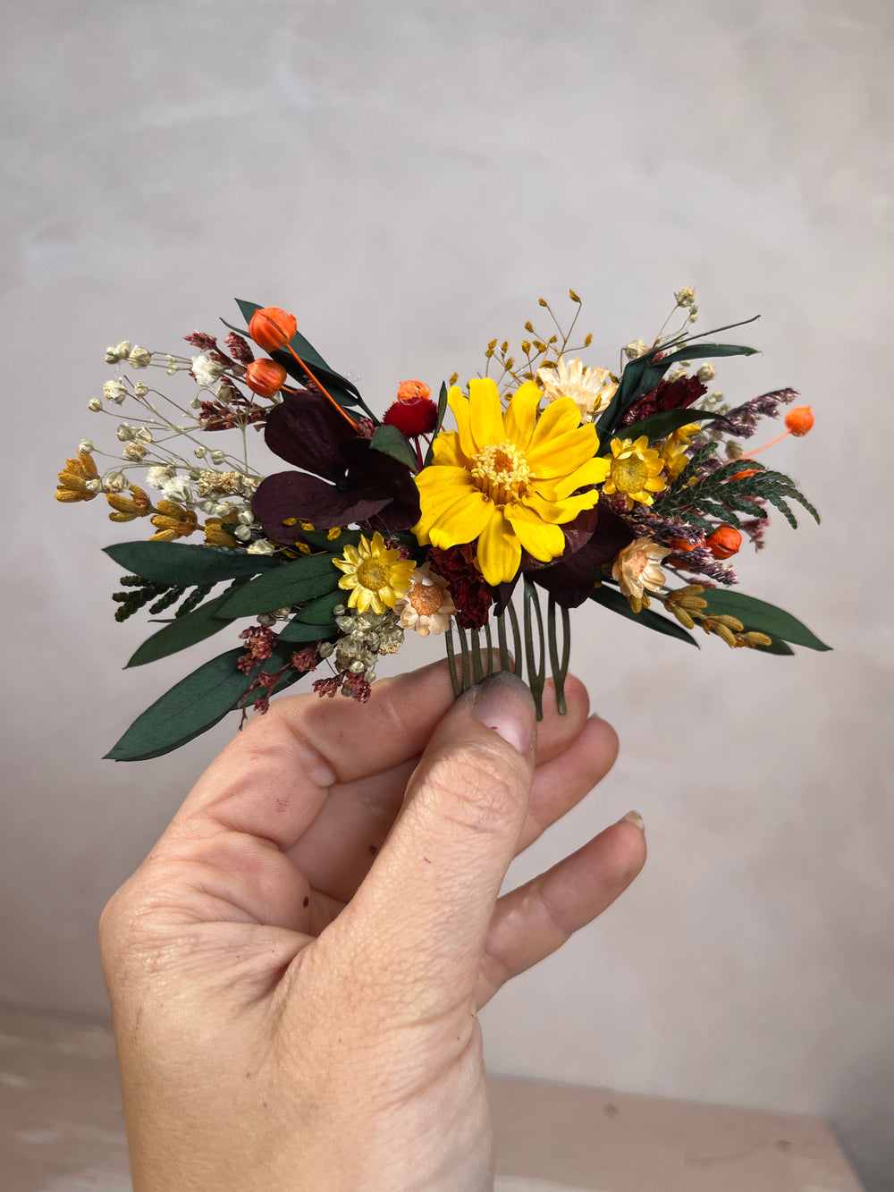 A hand holding a small dried flower hair comb featuring a prominent yellow cosmos flower, green foliage, and orange and burgundy accents against a neutral gray background.