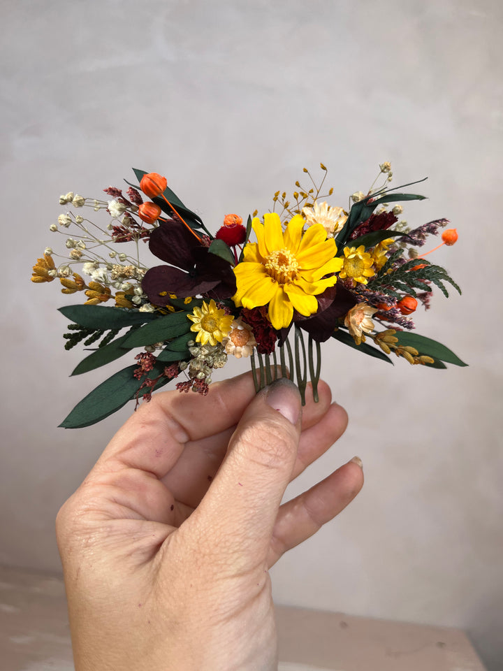 A hand holding a small dried flower hair comb featuring a prominent yellow cosmos flower, green foliage, and orange and burgundy accents against a neutral gray background.
