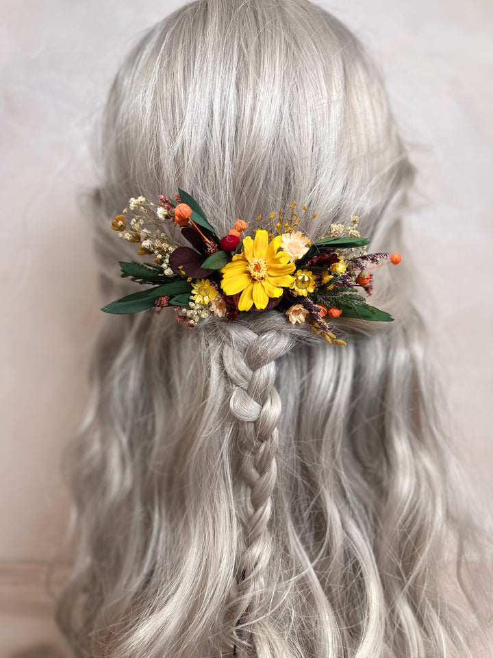 A long, wavy silver-gray wig with a braided low ponytail, styled with a small dried floral hair accessory featuring yellow flowers and green leaves, shown against a plain light beige background.