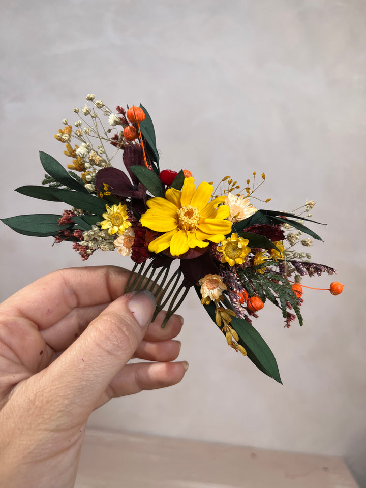 A hand holding a small dried floral hair comb accessory featuring a prominent yellow flower, green leaves, orange berries, and cream filler flowers against a plain light gray background.