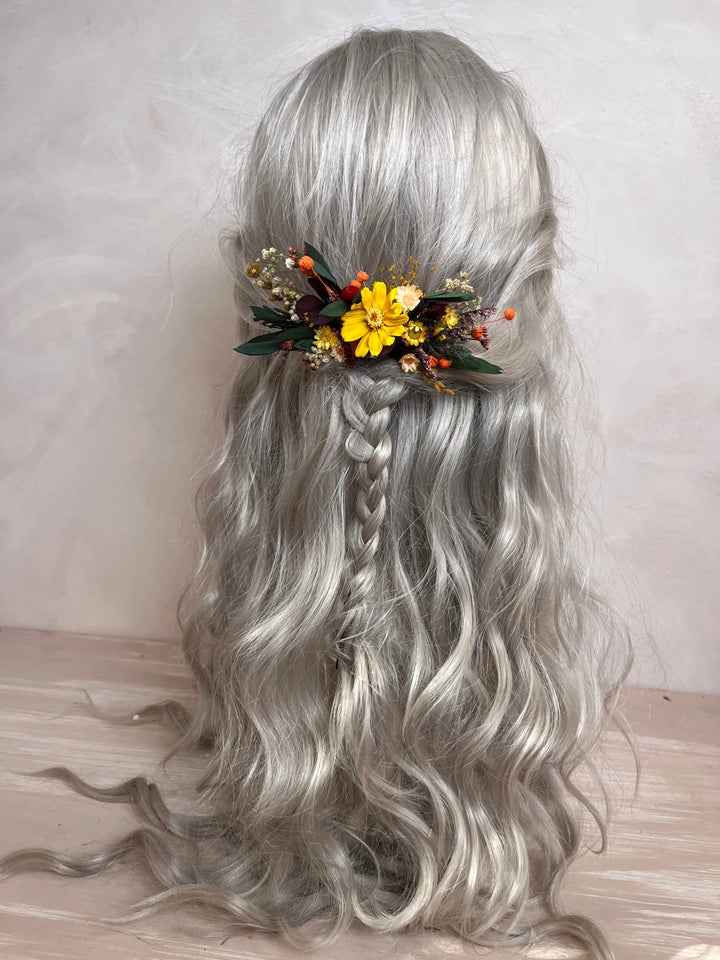 Long wavy silver-gray wig with a side braid and a floral hair accessory featuring yellow flowers and green leaves, displayed on a mannequin head against a neutral light background.