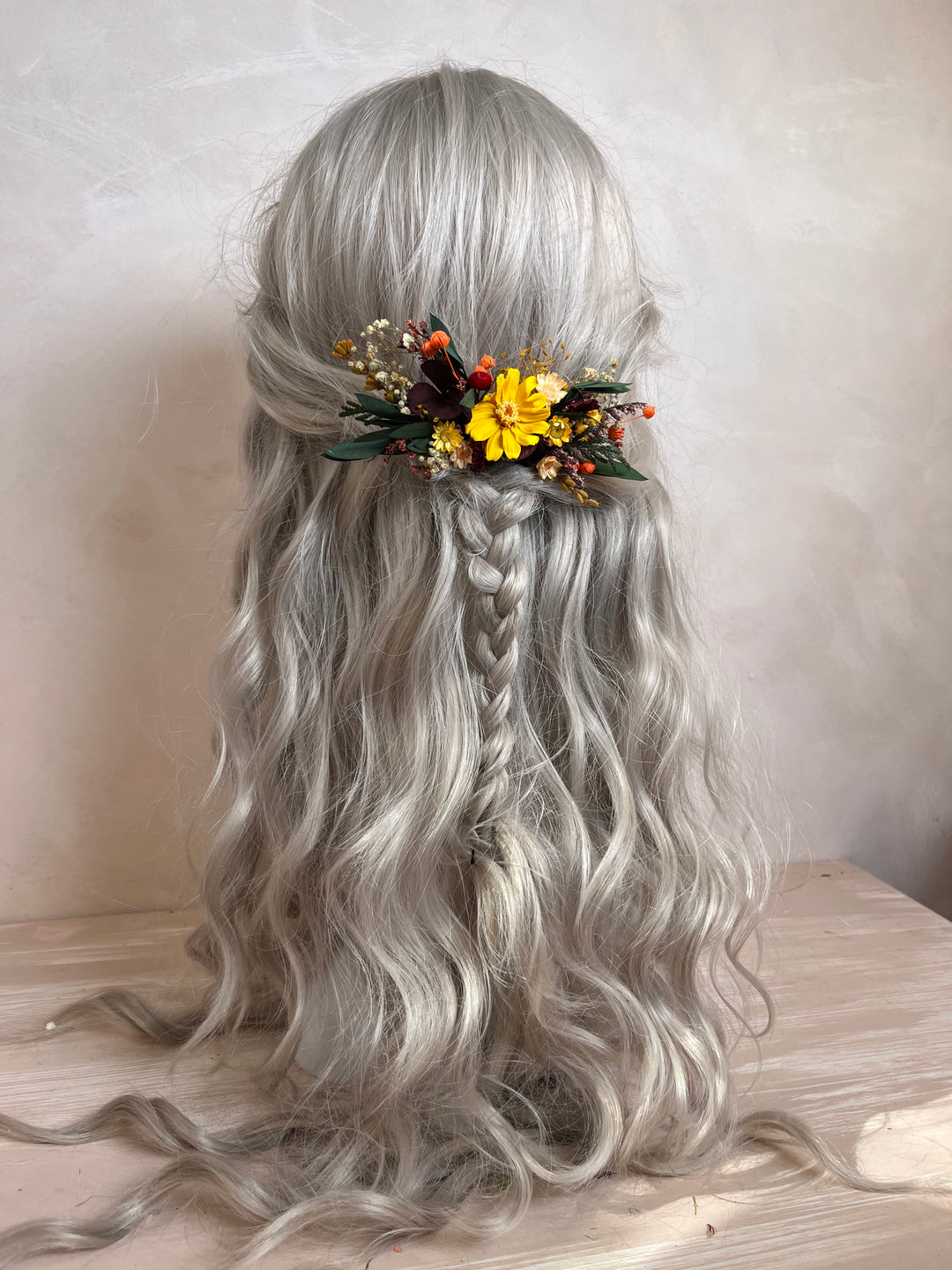 Long wavy silver-gray synthetic hair with a half-up style, a central braided section, and a decorative floral hair accessory with yellow flowers and green leaves, displayed on a mannequin head against a neutral beige background.