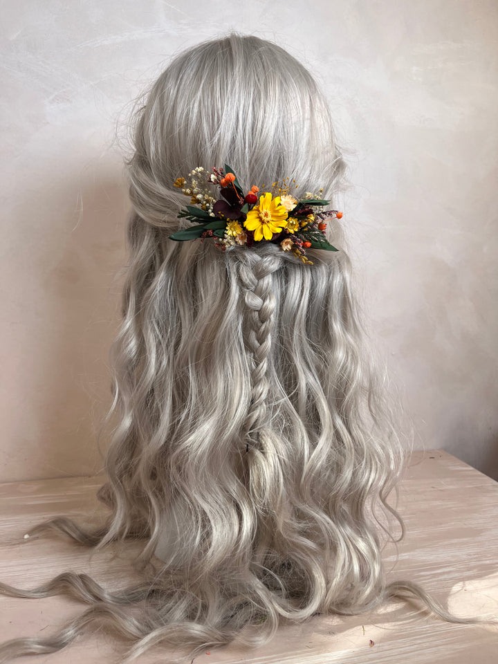 Long wavy silver-gray synthetic hair with a half-up style, a central braided section, and a decorative floral hair accessory with yellow flowers and green leaves, displayed on a mannequin head against a neutral beige background.