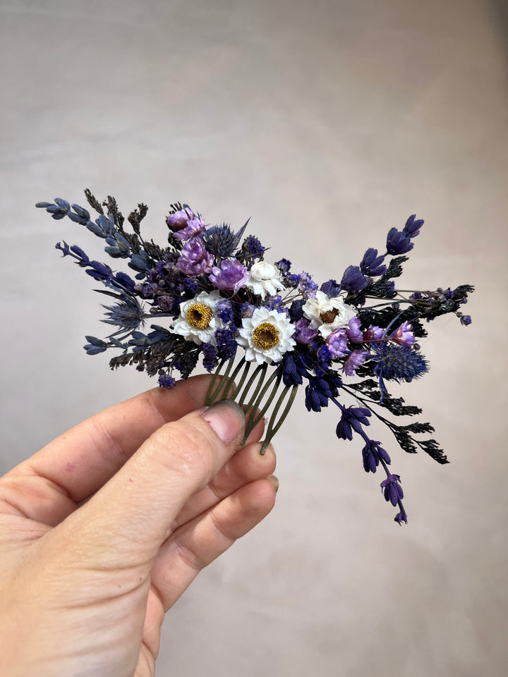 A hand holding a dried flower hair comb featuring purple lavender sprigs, small white daisies with yellow centers, and dark blue-gray filler flowers against a plain beige background.