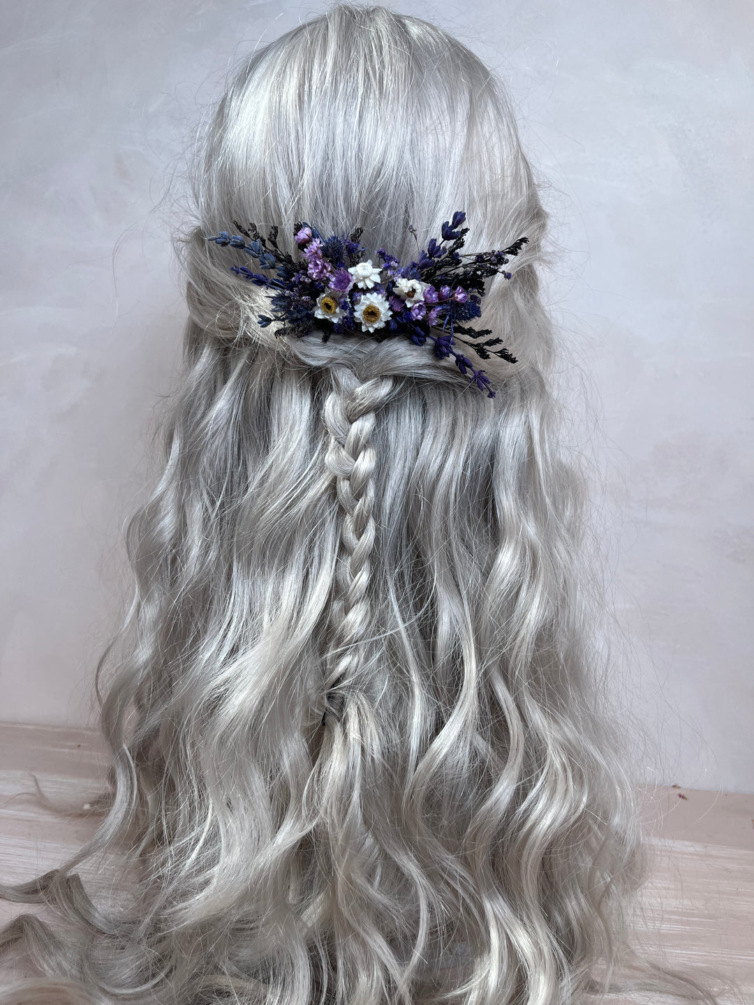 Long wavy silver-gray wig with a side braid and a floral hair accessory featuring purple, white, and yellow flowers, shown from the back against a plain light background.