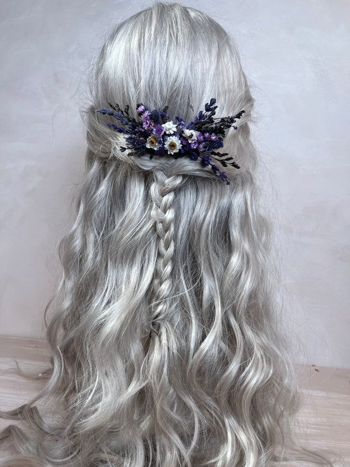 Long wavy silver-gray wig with a side braid and a floral hair accessory featuring purple, white, and yellow flowers, shown from the back against a plain light background.