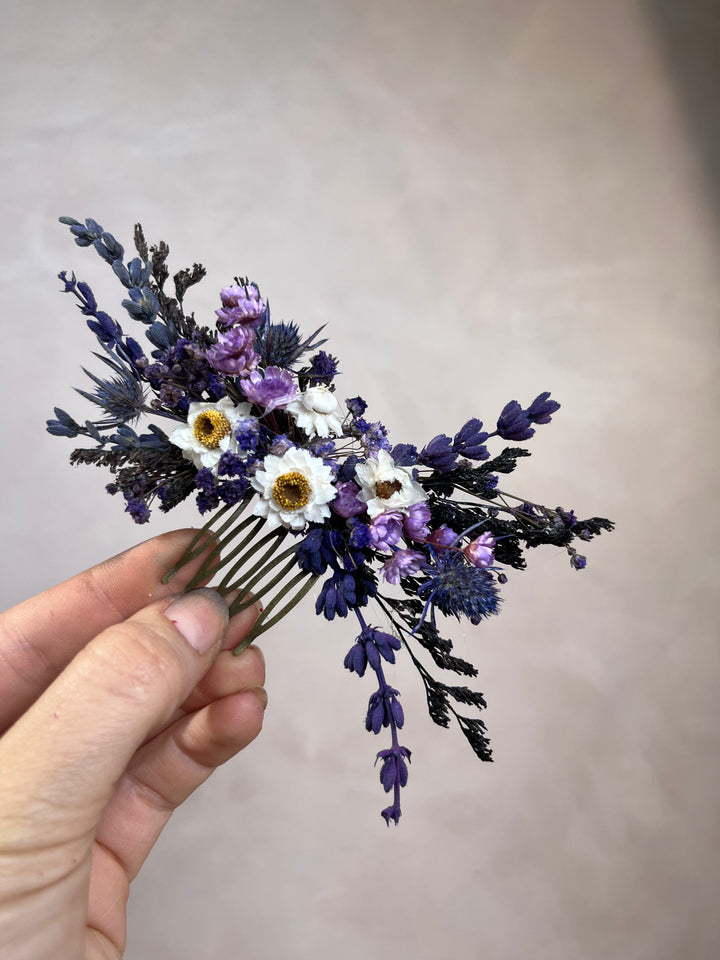 A hand holding a dried floral hair comb featuring lavender, purple wildflowers, and white daisies with yellow centers, set against a plain beige background.