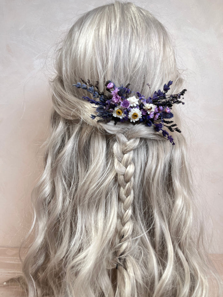 A long, wavy platinum blonde wig with a side braid and a decorative floral hair accessory in purple, white, and yellow, shown against a soft beige background.