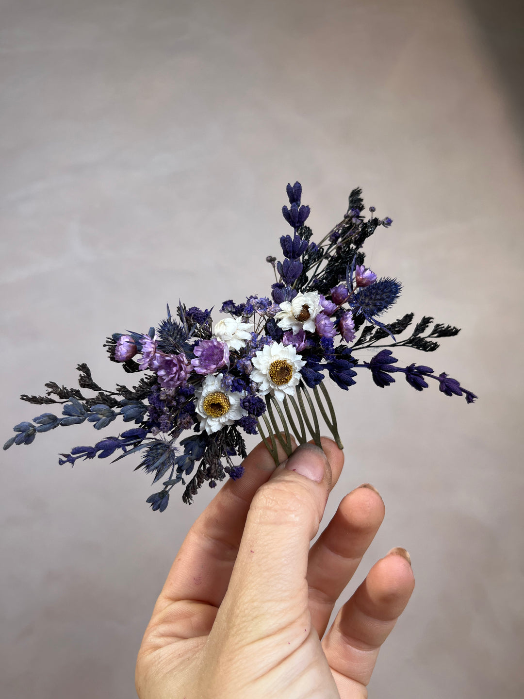 A hand holding a dried flower hair comb featuring purple lavender, white daisies, and blue thistles on a neutral gray background.