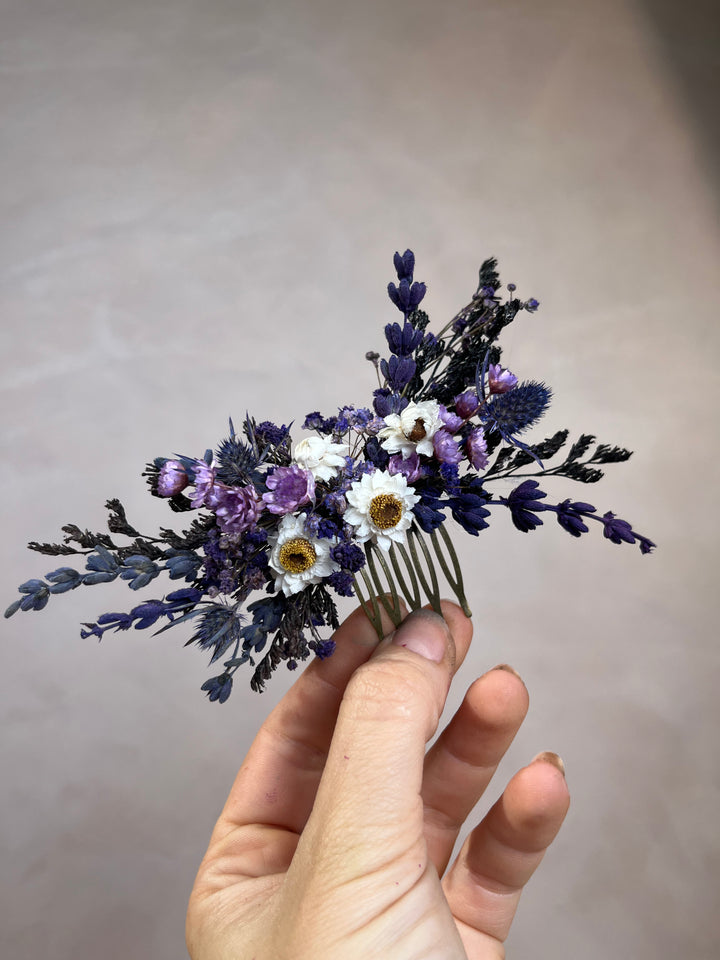 A hand holding a dried flower hair comb featuring purple lavender, white daisies, and blue thistles on a neutral gray background.