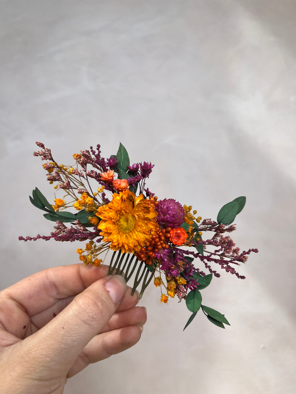 A hand holding a small dried flower hair comb accessory featuring orange, purple, and yellow flowers with green leaves against a neutral light gray background.