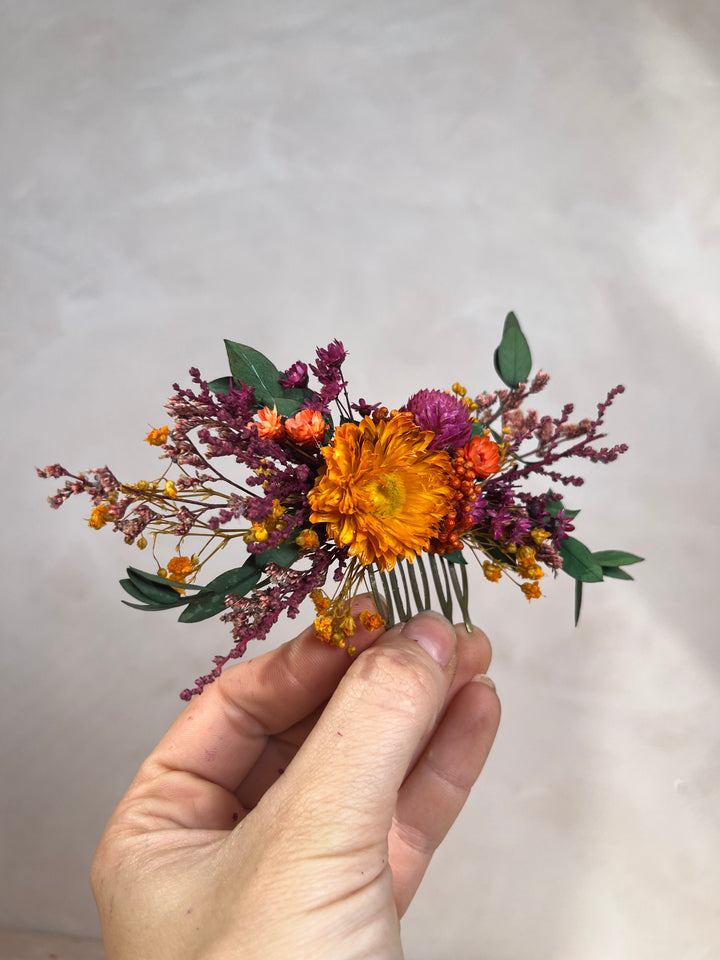 A hand holding a small dried flower hair comb featuring a prominent orange flower, purple blossoms, green leaves, and tiny yellow accents against a neutral light gray background.