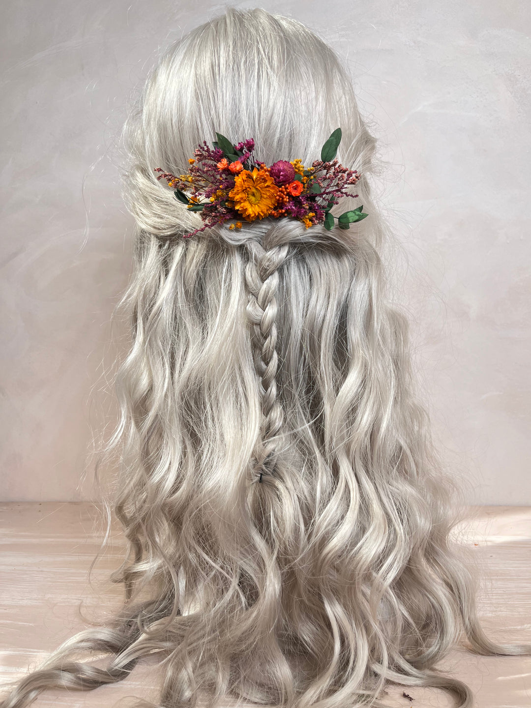 Long wavy silver-gray wig with a half-up style, center braid, and a floral hair accessory with orange, purple, and green flowers, shown from the back against a soft neutral background.