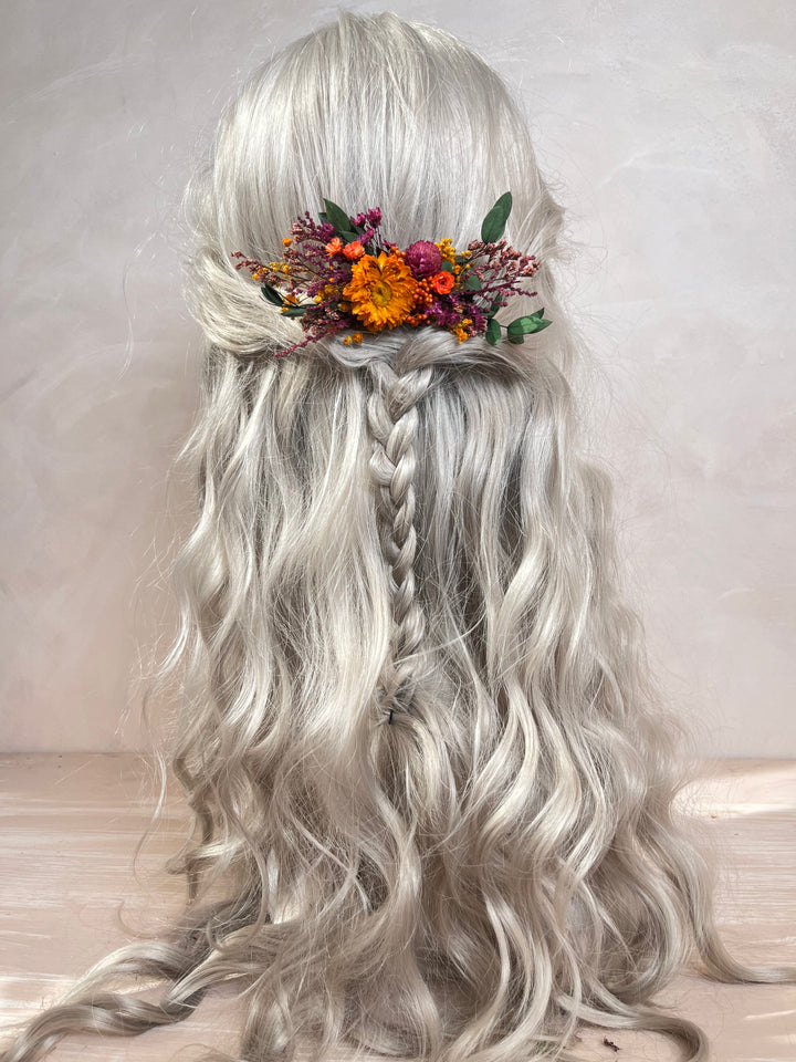 Long wavy silver-gray wig with a half-up style, center braid, and a floral hair accessory with orange, purple, and green flowers, shown from the back against a soft neutral background.