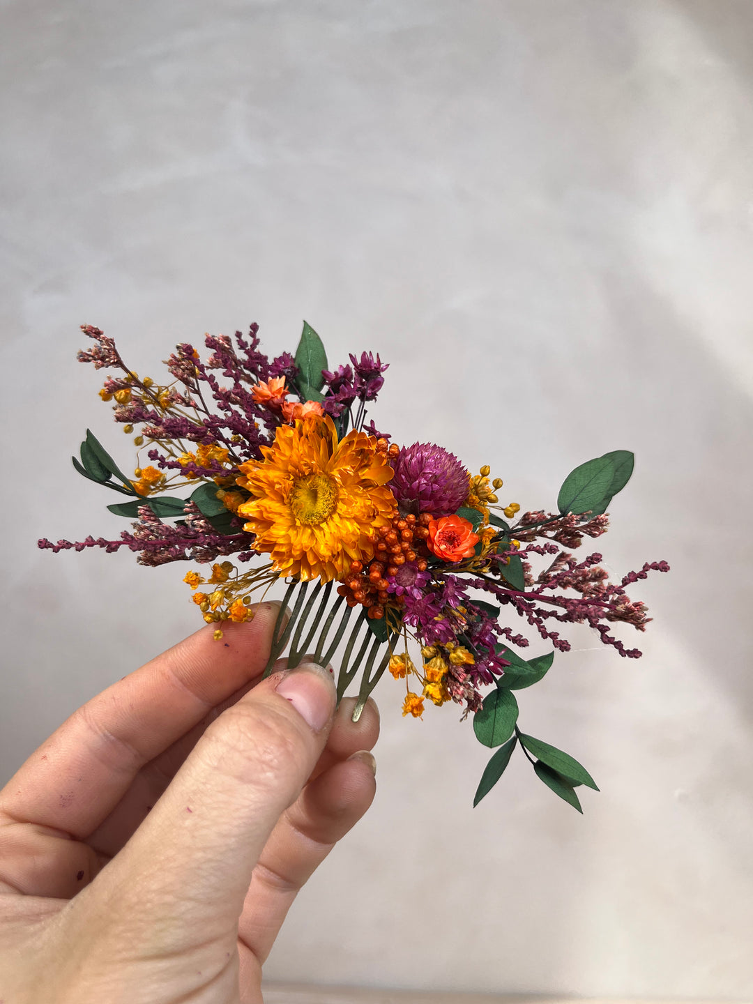 A hand holding a small dried flower hair comb featuring a bright orange flower, purple blooms, green leaves, and yellow accents against a neutral light gray background.