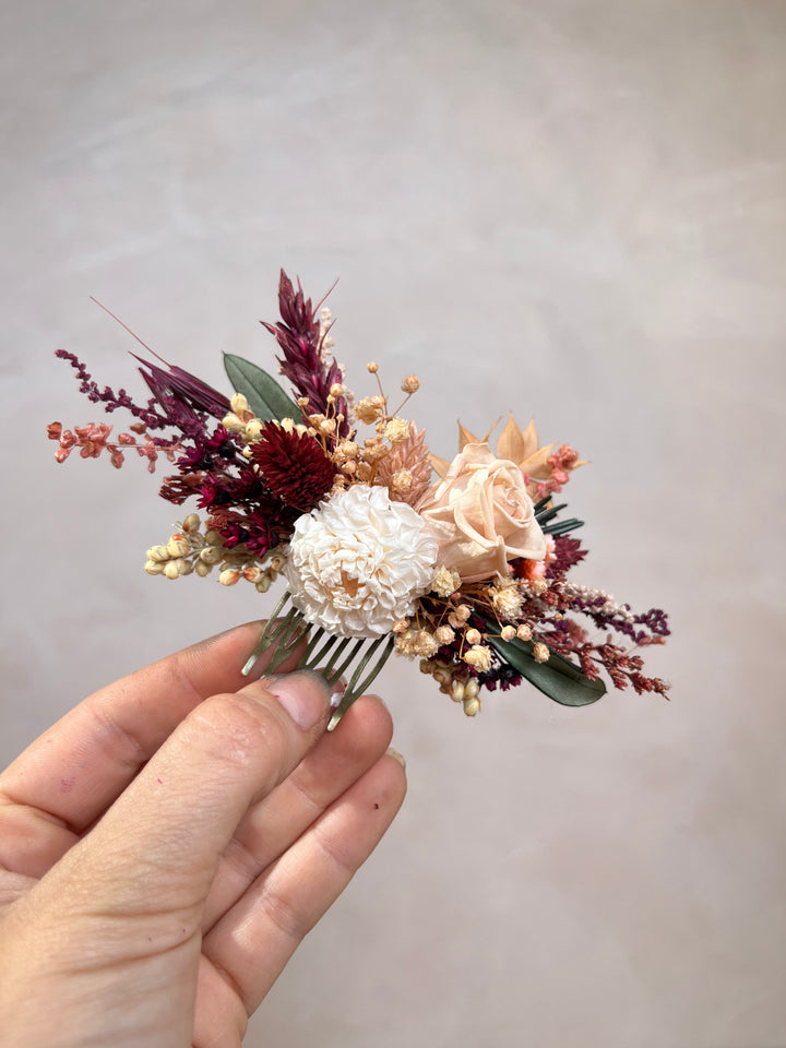 A dried flower hair comb accessory held by a hand, featuring cream, burgundy, and peach dried flowers with green leaves on a metallic comb base, set against a neutral textured background.