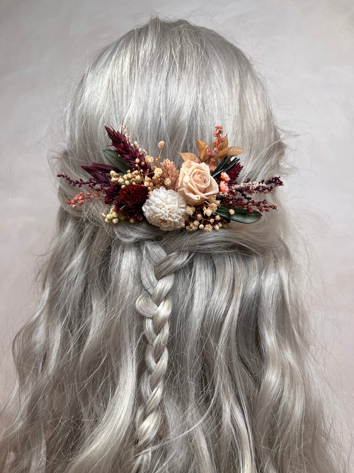 A long silver-gray wavy wig with a side braid, styled with a dried floral hair accessory in muted peach, burgundy, cream, and green tones, shown against a plain light background.