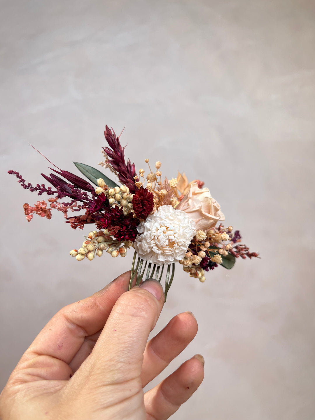 A hand holding a dried floral hair comb featuring cream dahlia, burgundy grasses, and beige filler flowers against a soft beige background.