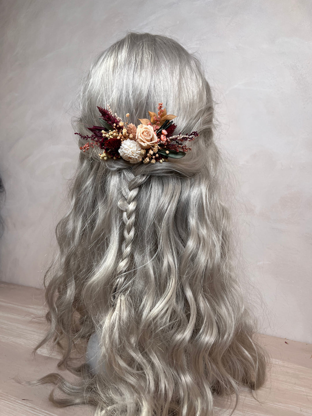 Long wavy silver-gray wig with a half-up style, braided side section, and a decorative floral hair accessory in muted peach, rust, cream, and burgundy tones, shown against a soft neutral wall.