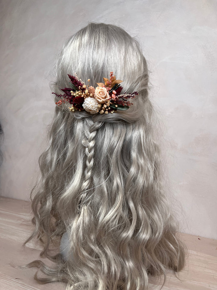 Long wavy silver-gray wig with a half-up style, braided side section, and a decorative floral hair accessory in muted peach, rust, cream, and burgundy tones, shown against a soft neutral wall.