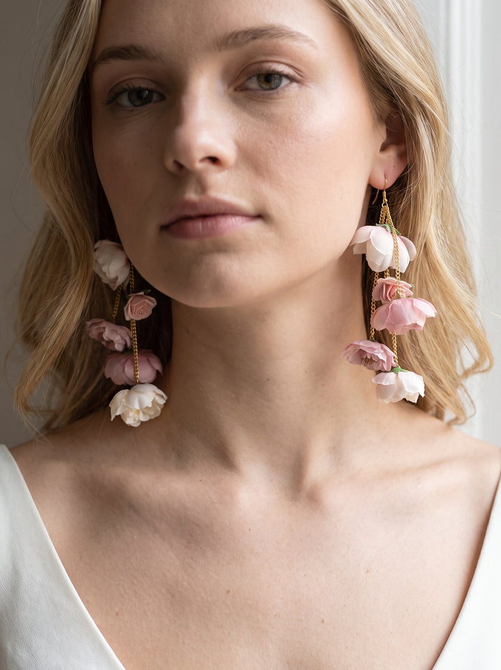 A close-up of a woman wearing large floral drop earrings with pink and white flower charms hanging from gold-toned hooks.