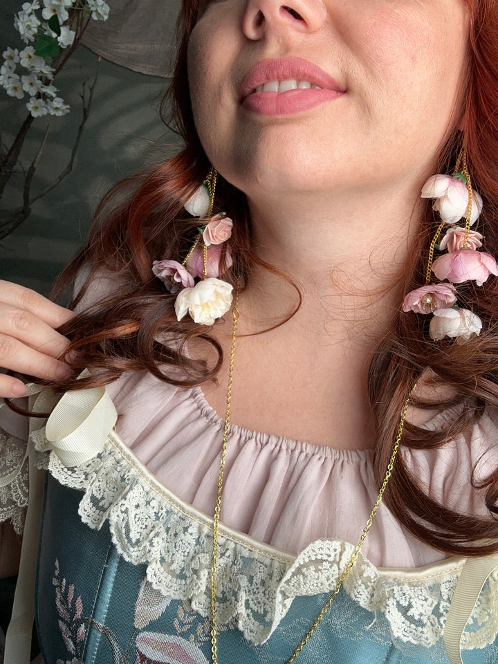 A close-up lifestyle shot of a woman wearing a delicate gold chain necklace adorned with small pink and white artificial flowers, styled in her hair over a light pink and blue floral dress.