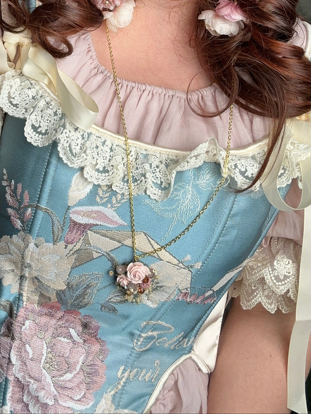 A woman wearing a detailed Lolita-style dress with a light blue satin bodice, pink ruffled sleeves, and white lace trim, styled with a floral headpiece and pink flower accessories.