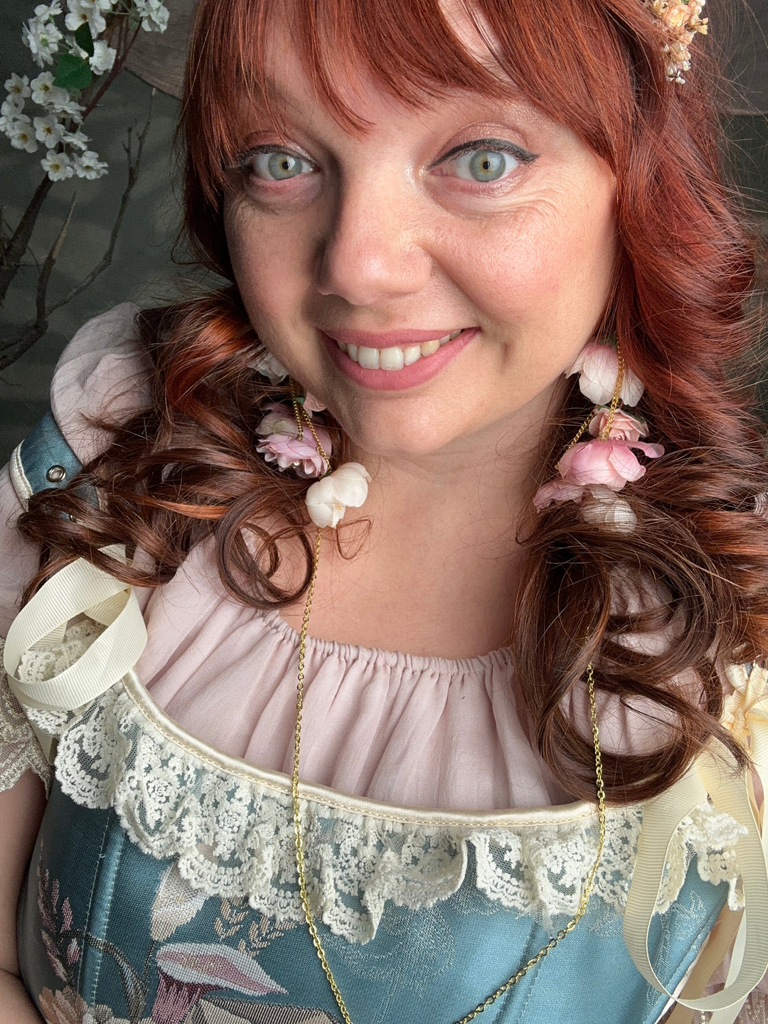 A close-up lifestyle shot of a woman with red hair wearing a vintage-style blue and pink dress with lace trim, floral hair accessories, and a gold chain necklace.