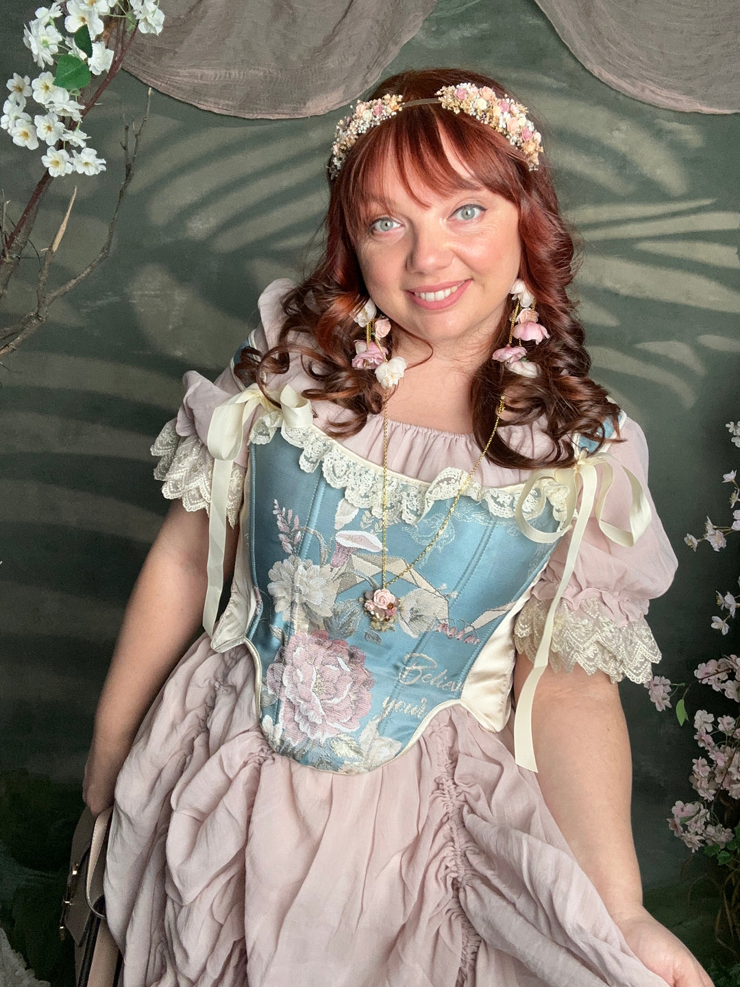 A woman wearing a vintage-style light blue and beige corset dress with floral embroidery, lace details, and a matching floral headpiece, photographed in a garden-like setting with cherry blossoms.