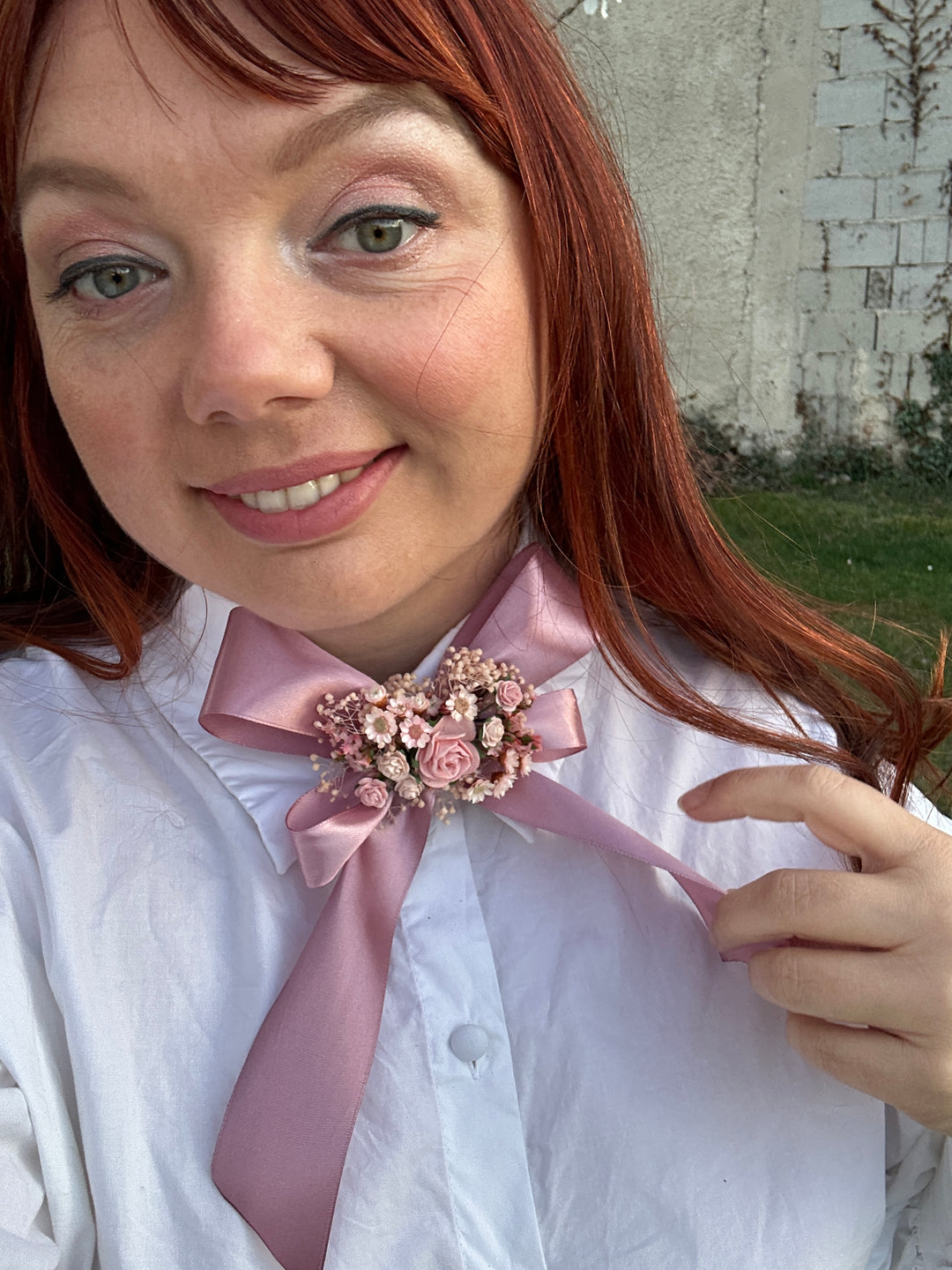 A close-up lifestyle shot of a smiling woman with red hair wearing a white button-up shirt and a dusty pink satin bow decorated with a floral brooch.