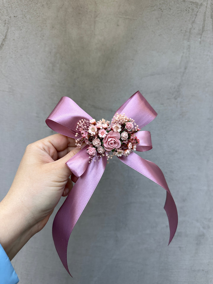 A hand holding a decorative pink ribbon and dried flower bouquet pin against a textured gray concrete background.