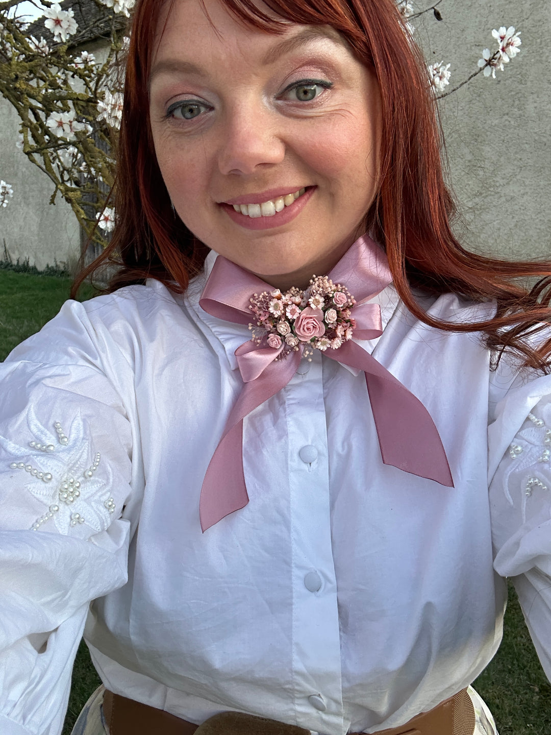 A smiling woman with red hair wearing a white long-sleeve button-up shirt with a dusty pink satin bow and a matching pink floral boutonniere on her chest, photographed outdoors.