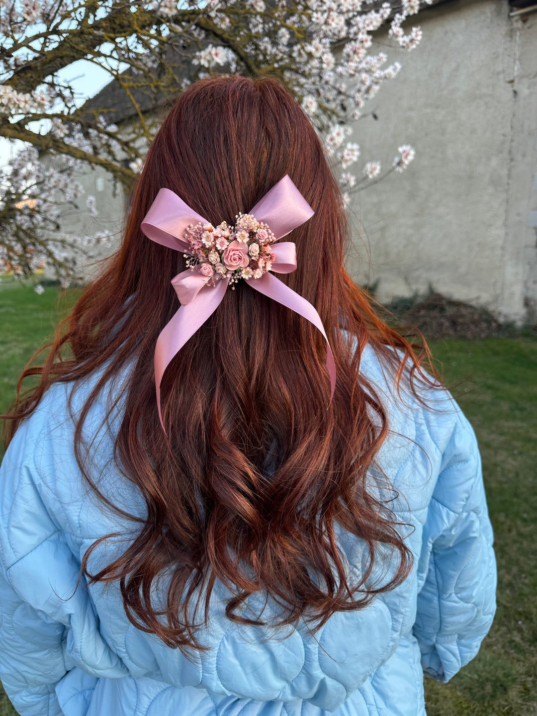 A long, wavy brown hair accessory with a light pink ribbon bow and a floral center, worn on a person's hair who is wearing a light blue puffer jacket outdoors.