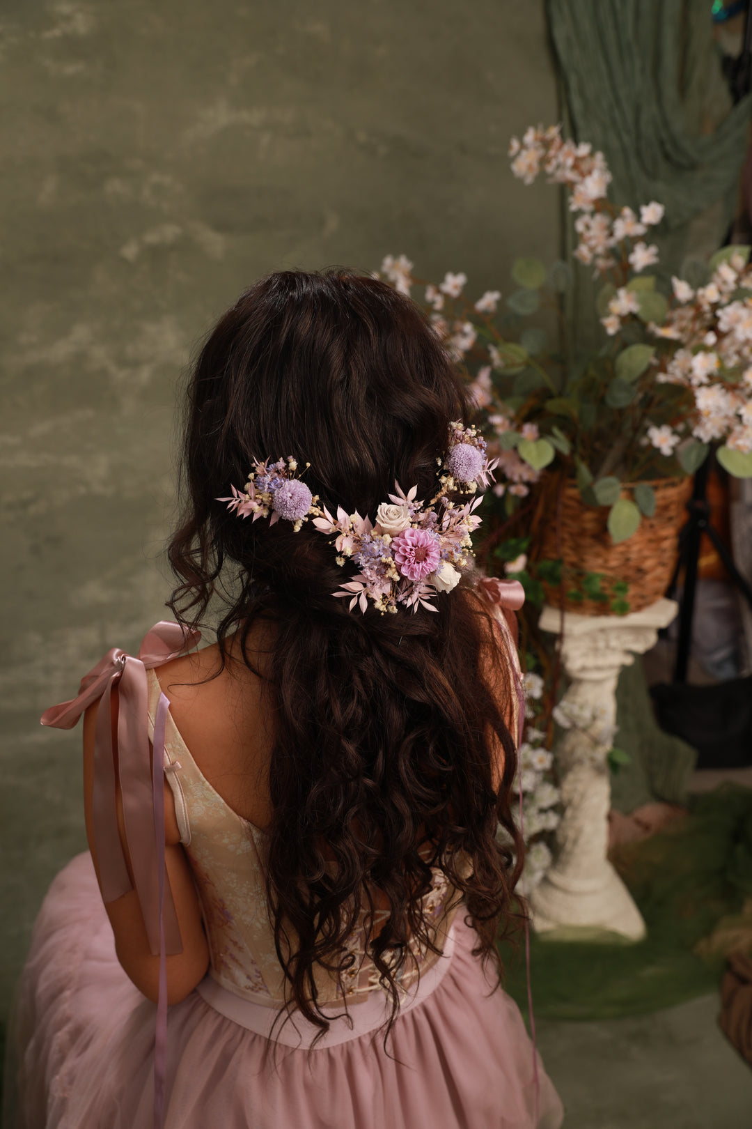 Woman with floral hair accessory in a pink dress standing in front of a green wall with plants.