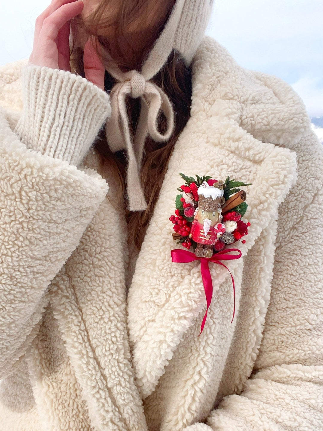  Woman in beige teddy coat with limited edition handmade Nutcracker Christmas brooch with red berries, pine cones, cinnamon and a bow for coats, hats and scarve