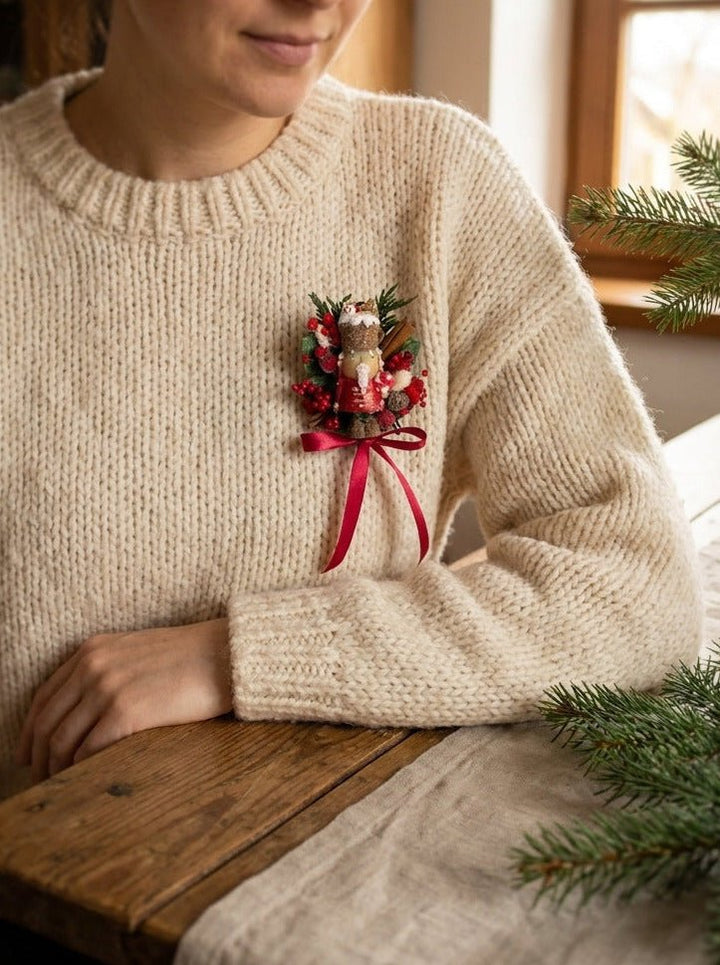 Woman in white sweater in cozy home with limited edition handmade Nutcracker Christmas brooch with red berries, pine cones, cinnamon and a bow for coats, hats and scarves