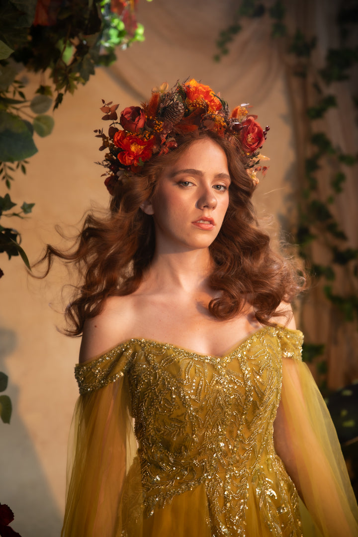 A woman wearing an ornate off-the-shoulder gold gown with intricate beadwork and a matching floral crown, posed against a warm beige backdrop with soft drapery and greenery.