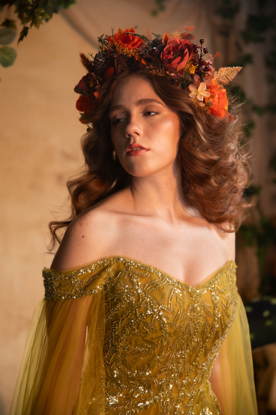 A woman wearing an off-the-shoulder gold gown with intricate beadwork and a matching floral crown of orange, red, and brown flowers, posed against a warm, softly lit background.