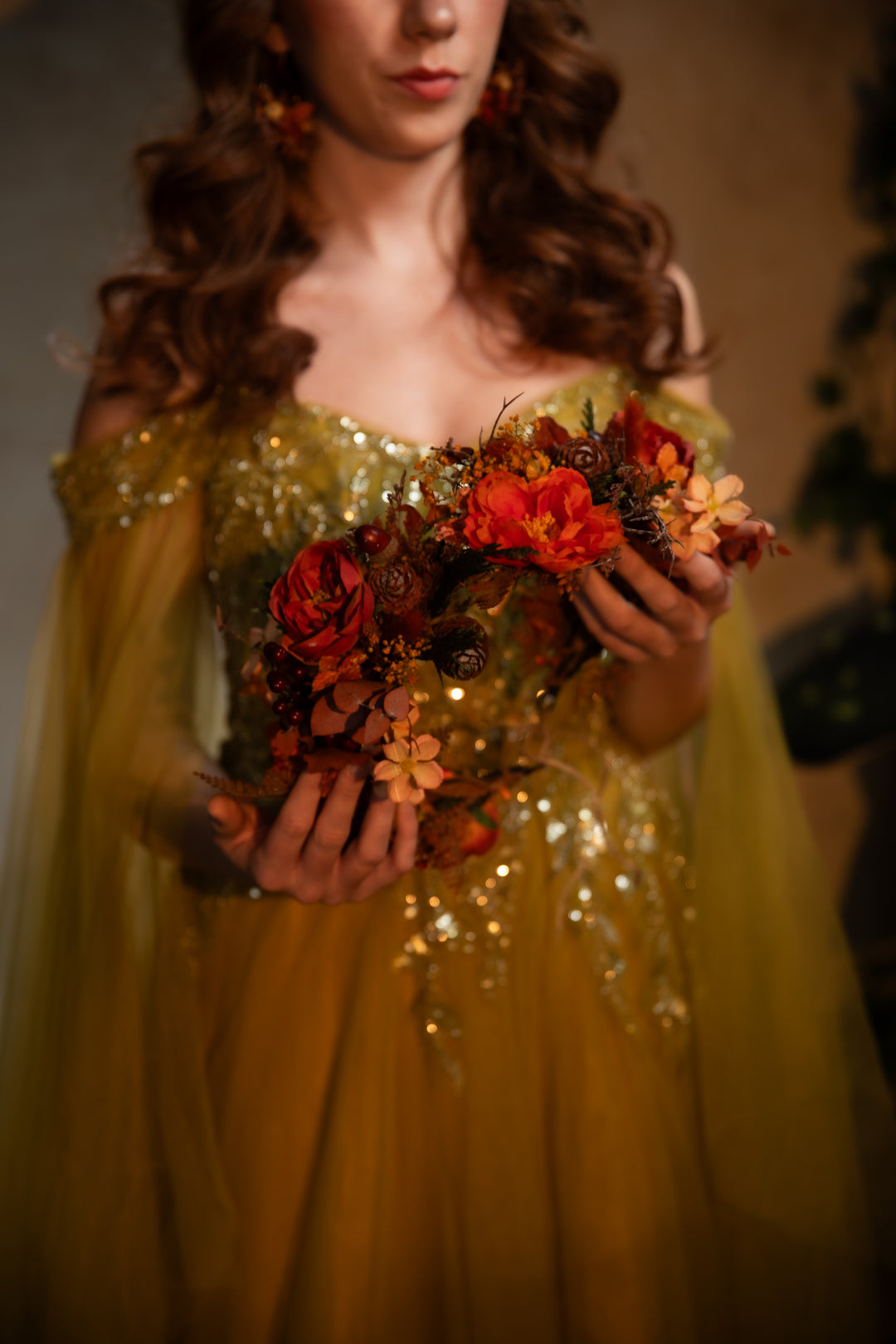 A woman in a shimmering gold off-the-shoulder gown holds a crown of red, orange, and brown autumn flowers against a warm, softly blurred background.