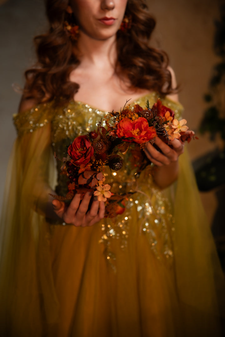 A woman in a shimmering gold off-the-shoulder gown holds a crown of red, orange, and brown autumn flowers against a warm, softly blurred background.