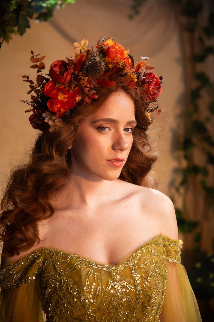 A woman with long wavy brown hair wearing a large floral crown of red, orange, and brown flowers, paired with an off-the-shoulder gold embellished gown.