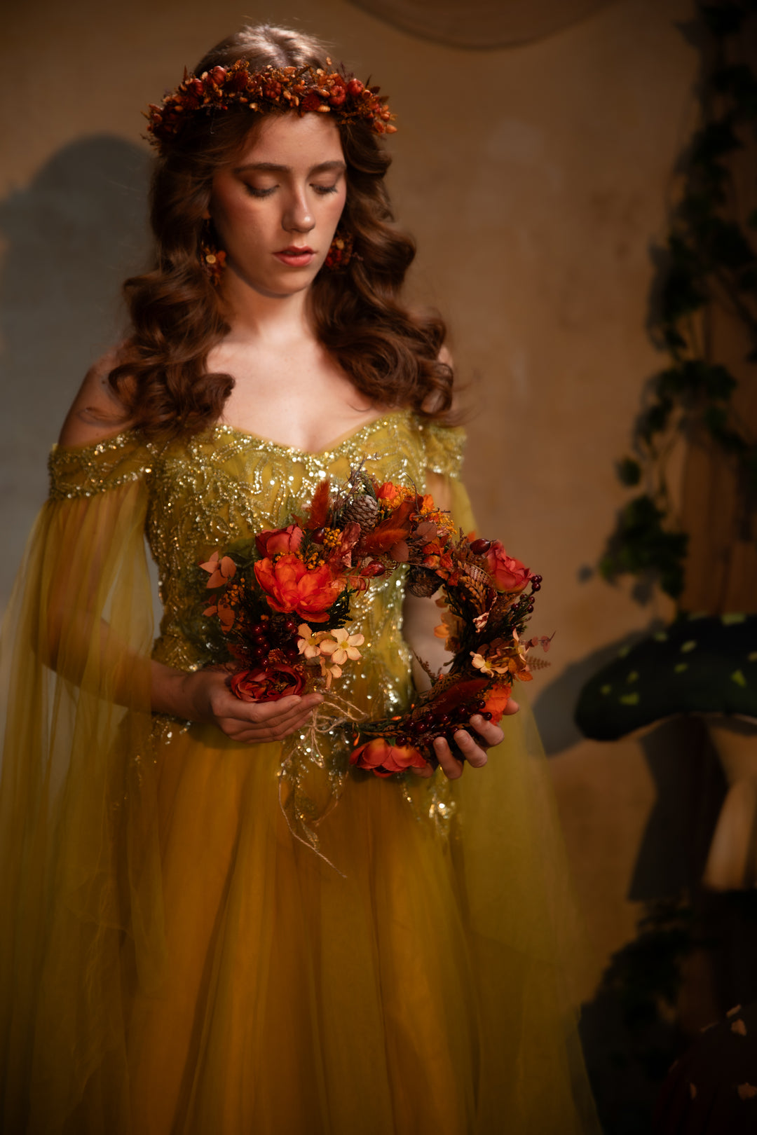 A woman in a golden green off-the-shoulder tulle gown with sequined detailing holds a floral wreath with red and orange autumn flowers. She wears a matching floral crown, set against a warm, dimly lit background with a mushroom prop on the right.