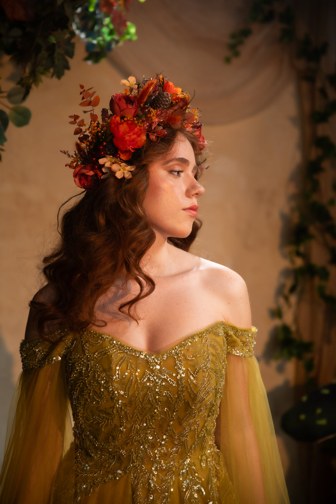 A woman in profile wearing an off-the-shoulder gold sequined gown with sheer tulle sleeves and a floral crown of red, orange, and brown flowers, posed against a warm beige backdrop with greenery.