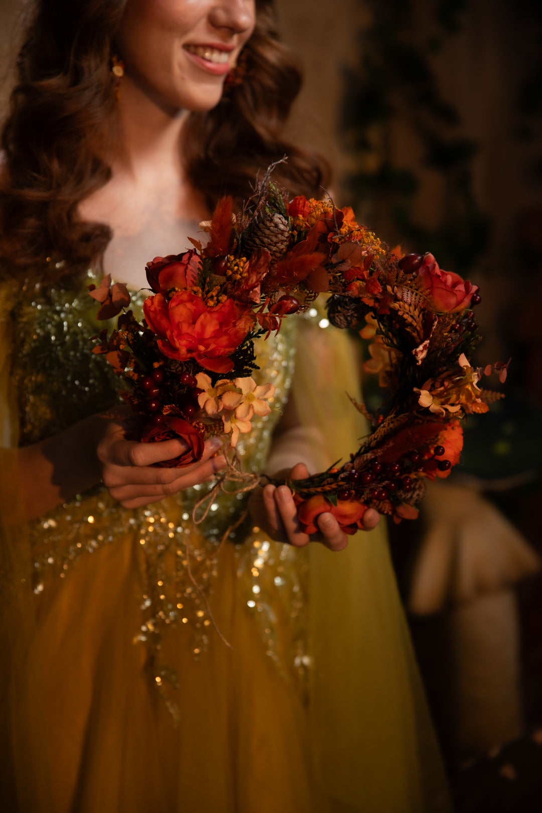 A woman in a sparkling gown holds a decorative autumn floral wreath with red flowers, pinecones, and dried foliage in a dimly lit indoor setting.