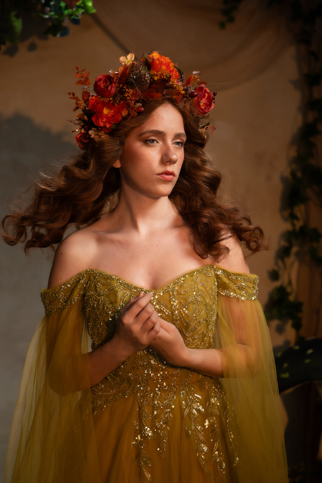A woman wearing an off-the-shoulder golden green gown with intricate gold embroidery and sheer tulle sleeves, styled with a vibrant orange and red floral crown. She is posed against a warm, softly lit, rustic background.