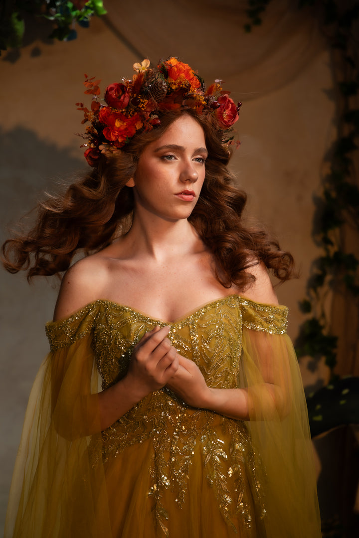 A woman wearing an off-the-shoulder golden green gown with intricate gold embroidery and sheer tulle sleeves, styled with a vibrant orange and red floral crown. She is posed against a warm, softly lit, rustic background.